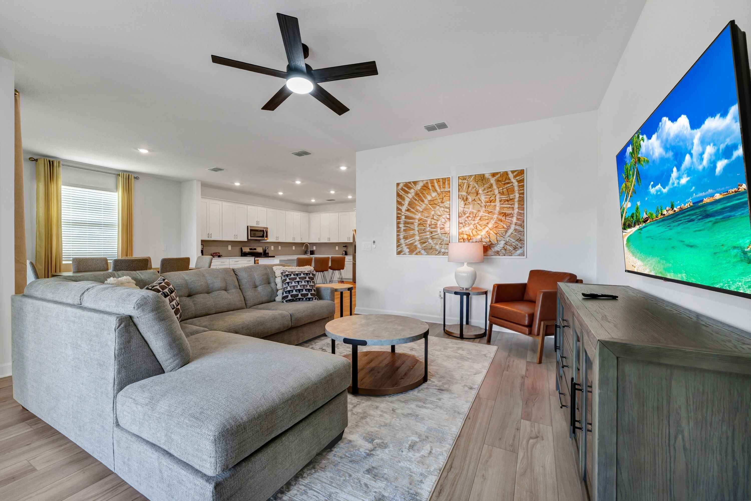 A modern living room with a sectional sofa, a coffee table, and a TV. There is a kitchen in the background with white cabinets and a ceiling fan.