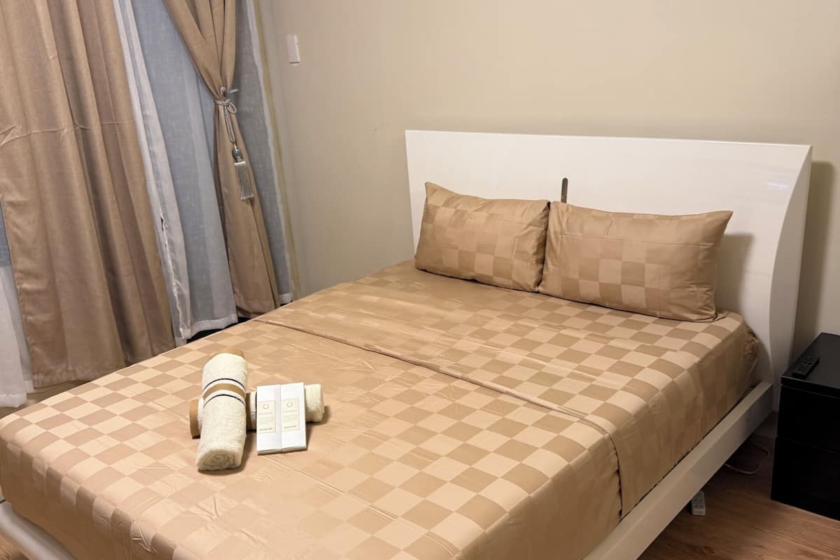 A neatly made bed with a checkered beige bedspread, two pillows, and a rolled towel.