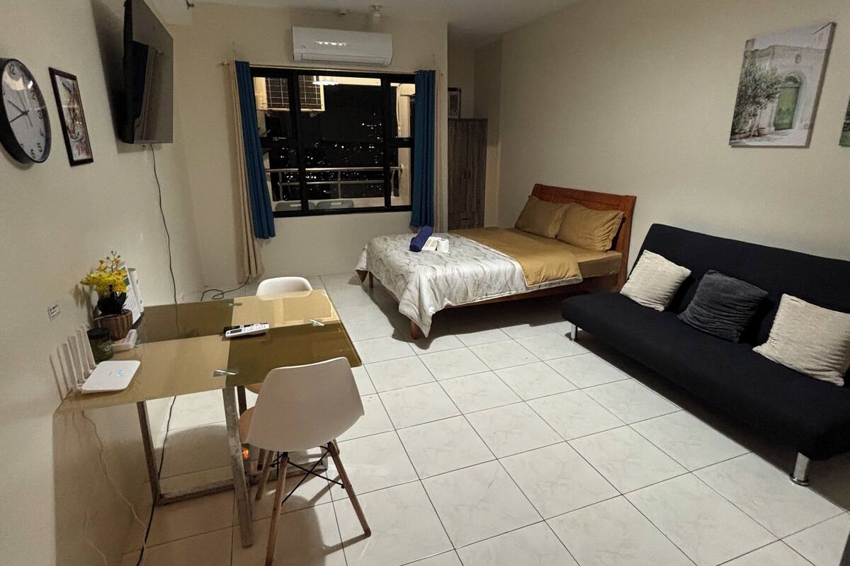 A room with a bed, a sofa, a dining table with chairs, a TV mounted on the wall, and a window with curtains.