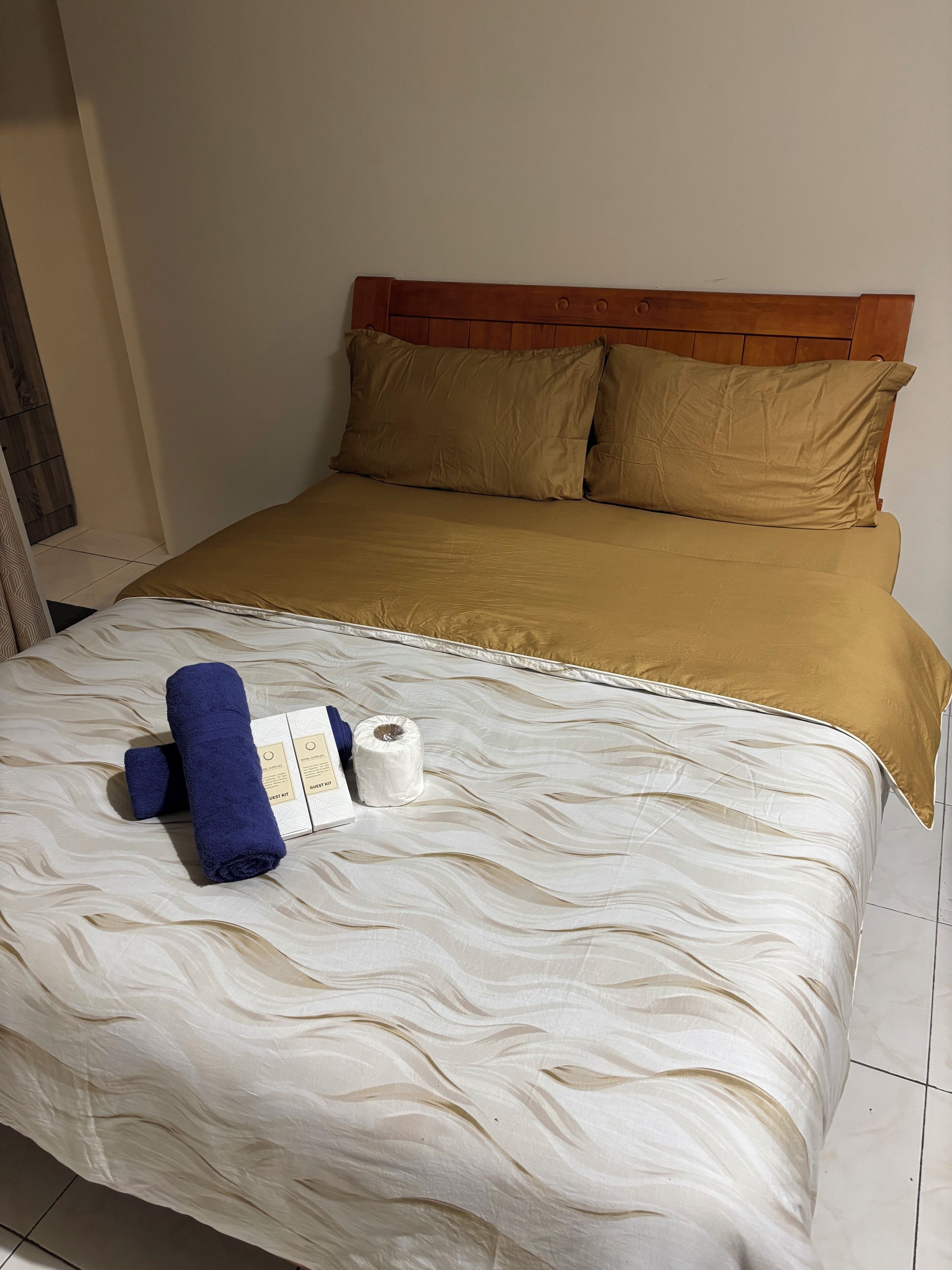 Una cama bien hecha con cabecera de madera, colcha color beige y marrón, una toalla azul y un paquete blanco sobre la cama.