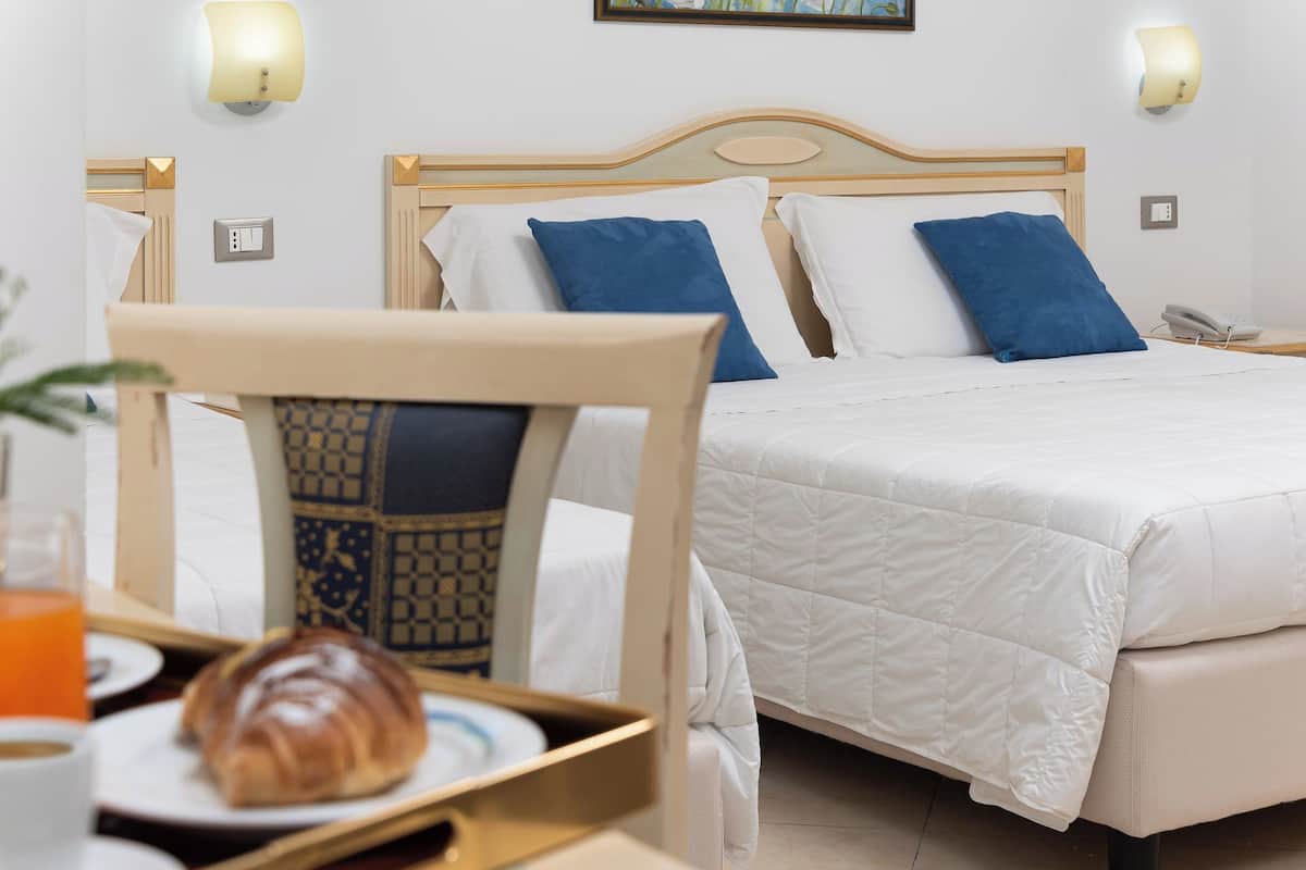 A hotel room with a bed, two bedside lamps, a bedside phone, and a chair with a tray table.