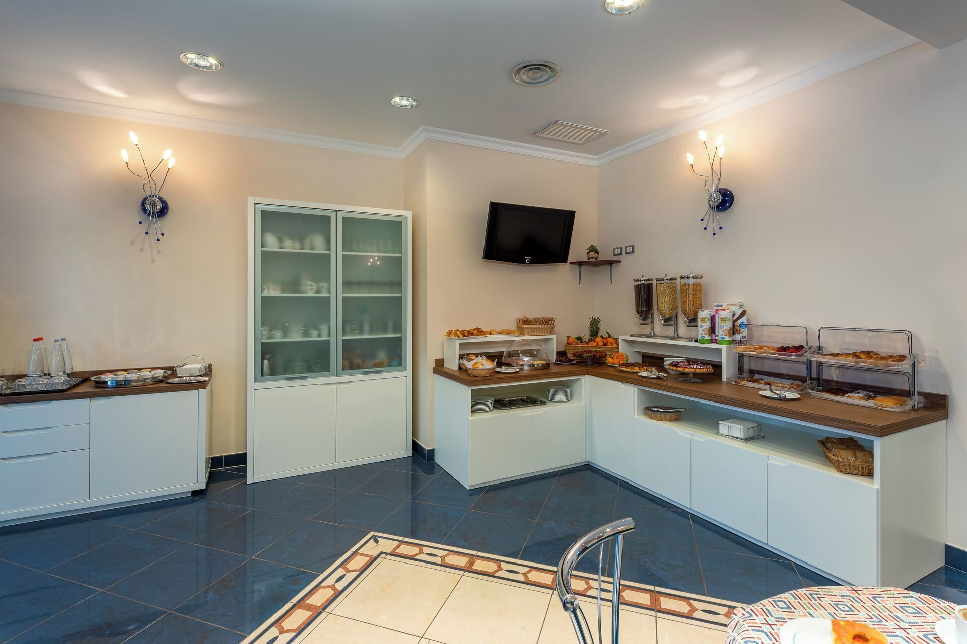 Une cuisine moderne équipée d’un réfrigérateur encastré, d’un four à micro-ondes et d’une machine à café. On y trouve un buffet garni de divers aliments, ainsi qu’un téléviseur fixé au mur.