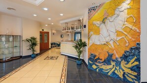 A hotel lobby with a tiled floor, a reception desk, a glass cabinet with decorative items, and a large mural with horses. - Helios Hotel (San Vito Lo Capo)
