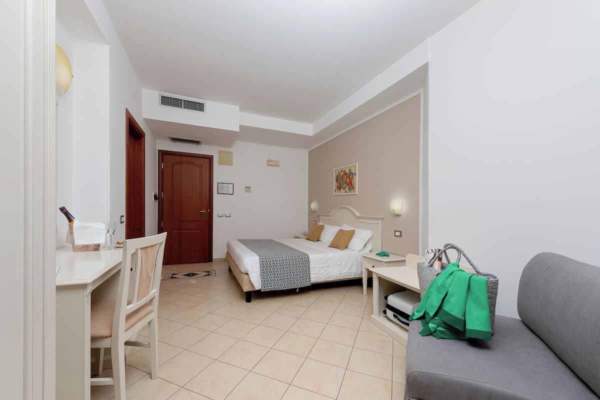 A hotel room with a bed, a desk, a chair, a sofa, and a small table with a green bag on it.