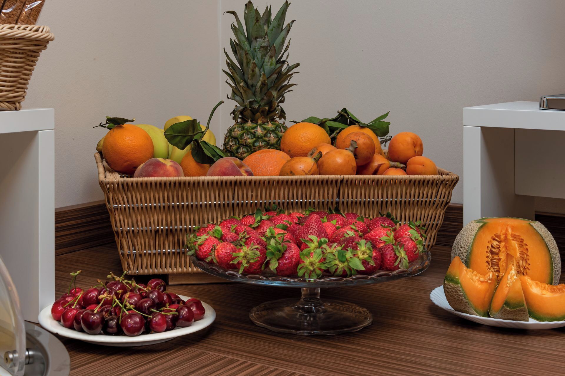 Une présentation de fruits comprenant un panier en osier, un présentoir à gâteaux en verre et une assiette de cerises.