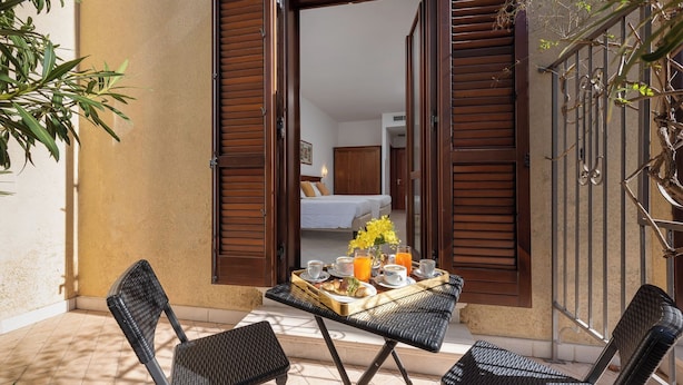 An open wooden door leading to a bedroom with a bed, a dining table with breakfast items, and two wicker chairs. - Helios Hotel (San Vito Lo Capo)