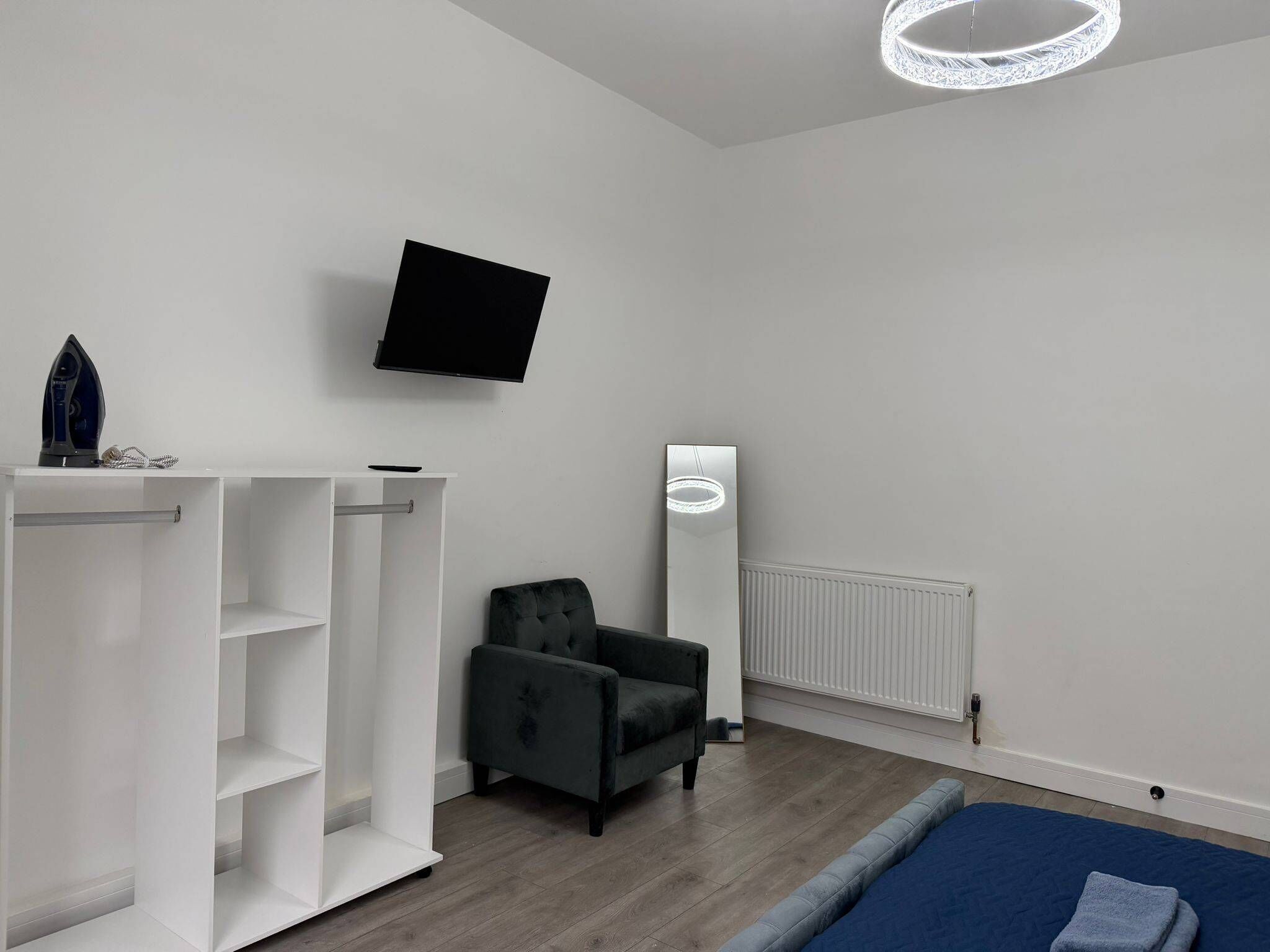 A modern bedroom with a bed, a grey armchair, a white shelving unit, a flat-screen TV, and a floor lamp.