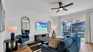 A modern living room with a sofa, a TV, a ceiling fan, and a view of a house through large windows. - Reunion Village Home near Disney: Private Pool & Waterpark - 7880ML (Kissimmee)