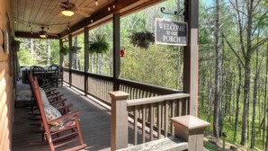 A wooden porch with a 'Welcome to our Porch' sign, outdoor seating, and a view of a forested area. - New! Rocco's creekside cabin, relax and wine down on the newly screened in porch (Ellijay)