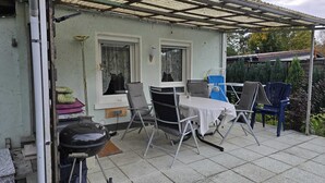 Een overdekt terras met een tafel, stoelen, een barbecue en een raam met kantgordijnen.