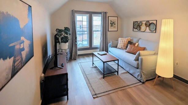 A cozy living room with a sofa, coffee table, and a TV on a stand. There is a large window with curtains, a plant, and artwork on the walls. - The Green House, RiNo - Luxury Queen Bed, Pullout Couch & Full Kitchenette (Denver)