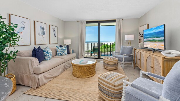 A living room with a sofa, armchairs, a coffee table, and a TV. There are framed pictures on the wall and a view of the ocean through the balcony doors. - Healthier Beachfront Stay - Short Walk to Flagler (New Smyrna Beach)