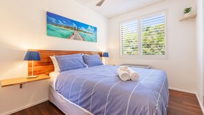 Quarto com cabeceira de madeira, cama com roupa de cama azul, duas luminárias de cabeceira, janela com persianas e uma pintura de uma praia.