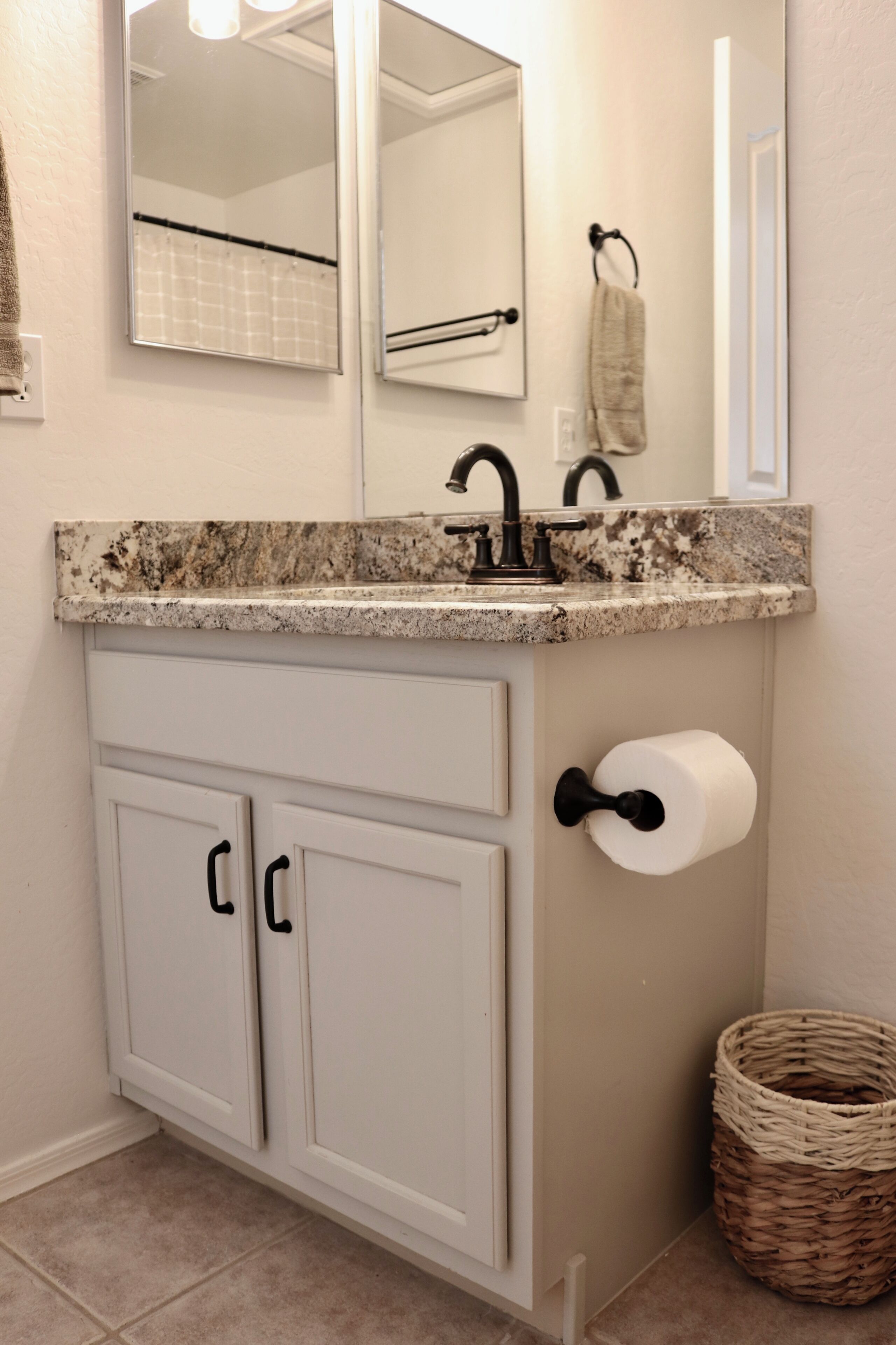 Un bagno con un lavabo in marmo, mobiletti bianchi, uno specchio, un portasciugamani e un cesto in vimini.