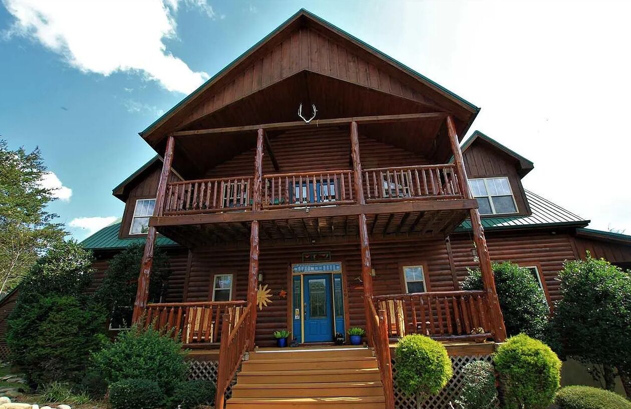 A two-story log cabin with a balcony, a blue door, and a deer head decoration above the entrance.