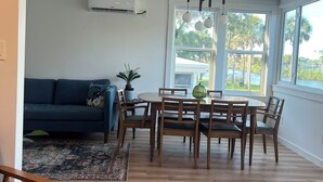 Uma sala de estar moderna com um sofá azul, mesa de jantar de madeira com cadeiras e uma grande janela com vista para palmeiras.