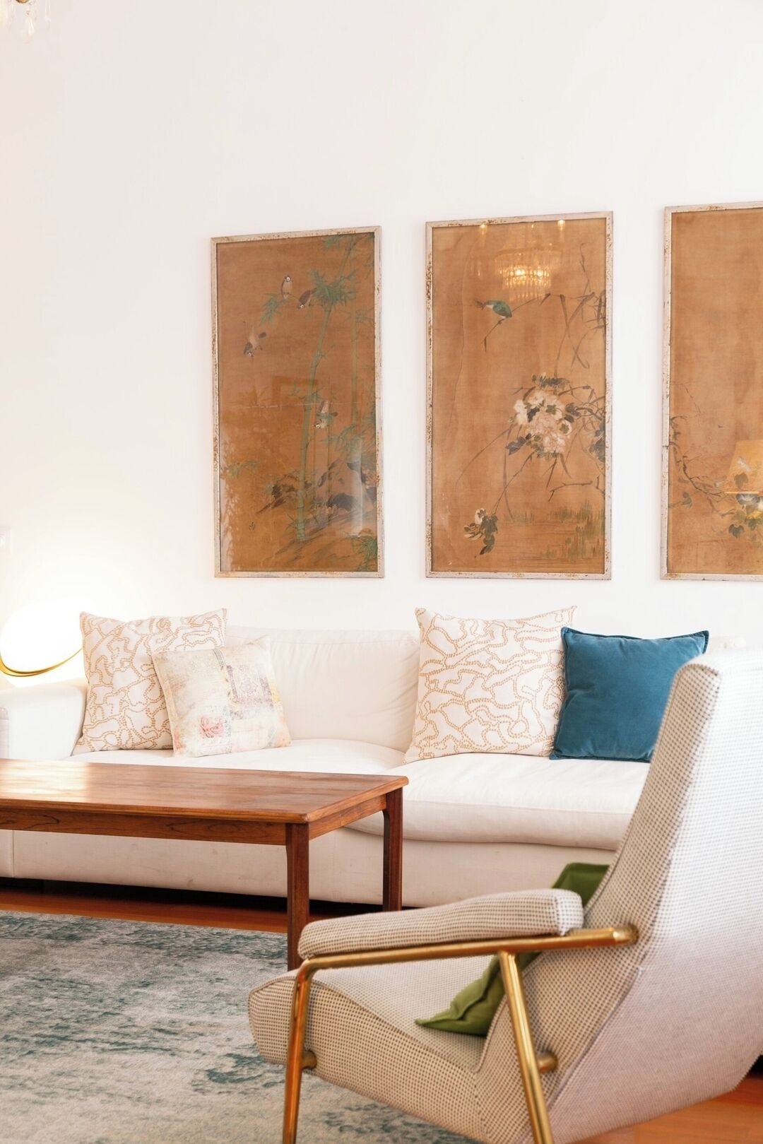 Un salon avec un canapé blanc, une table basse en bois et un fauteuil à motifs. Trois grandes peintures abstraites ornent le mur.