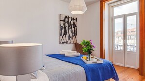 A hotel room with a bed, a bedside table with a lamp, a window with a view of buildings, and a painting on the wall. - Baixa most luminous 3 BDR flat!! Great WI-FI!!! (Lisboa)
