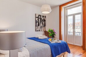 Ein Hotelzimmer mit einem Bett, einem Nachttisch mit Lampe, einem Fenster mit Blick auf Gebäude und einem Bild an der Wand.