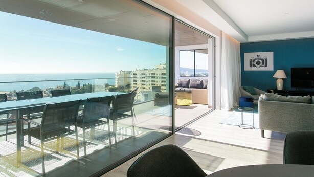 Et moderne hotelværelse med balkon med havudsigt, et spiseområde med et glasbord, og et opholdsområde med sofa og fjernsyn.
