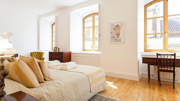 Un dormitorio con una cama, un escritorio de madera y una silla. Dos ventanas dejan entrar luz natural.