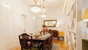 Un comedor con una mesa de madera dispuesta para cuatro personas, rodeada de sillas. Un candelabro cuelga del techo, y un espejo está colgado en la pared.