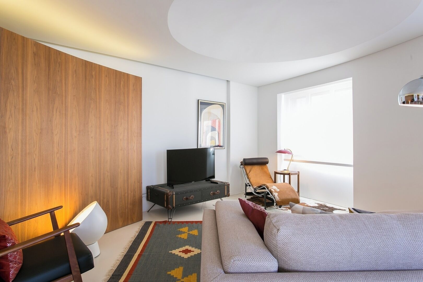 Un salon moderne comprenant une télévision, un fauteuil en cuir, un canapé, une petite table avec une lampe et une grande fenêtre laissant entrer la lumière naturelle.