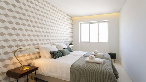 Ein modernes Schlafzimmer mit Wandpaneelen im geometrischen Muster, einem Bett mit weißen Bettwäsche und mehreren Kissen, einer Nachttischlampe und einem Fenster, das natürliches Licht hereinlässt.