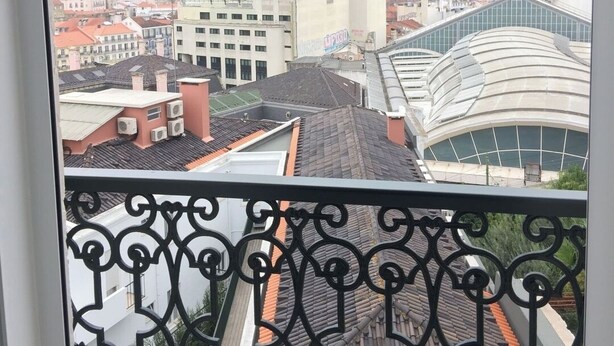 A cityscape view from a balcony with a wrought iron railing, featuring a mix of traditional and modern buildings, including a distinctive curved glass structure.