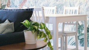 Een kamer met een eethoek, met witte stoelen en een tafel, een donkergrijze bank met kussens, en een potplant.