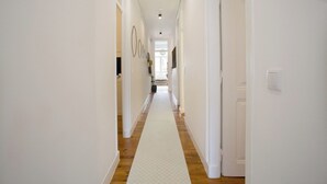 A hallway with white walls, wooden flooring, and a white carpet. There are doors on both sides, and a light switch is visible on the right wall.