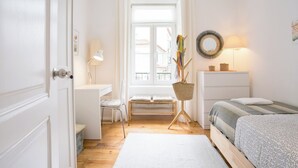 A bright, white room with a desk, chair, and bed. There is a window with a view of buildings outside.
