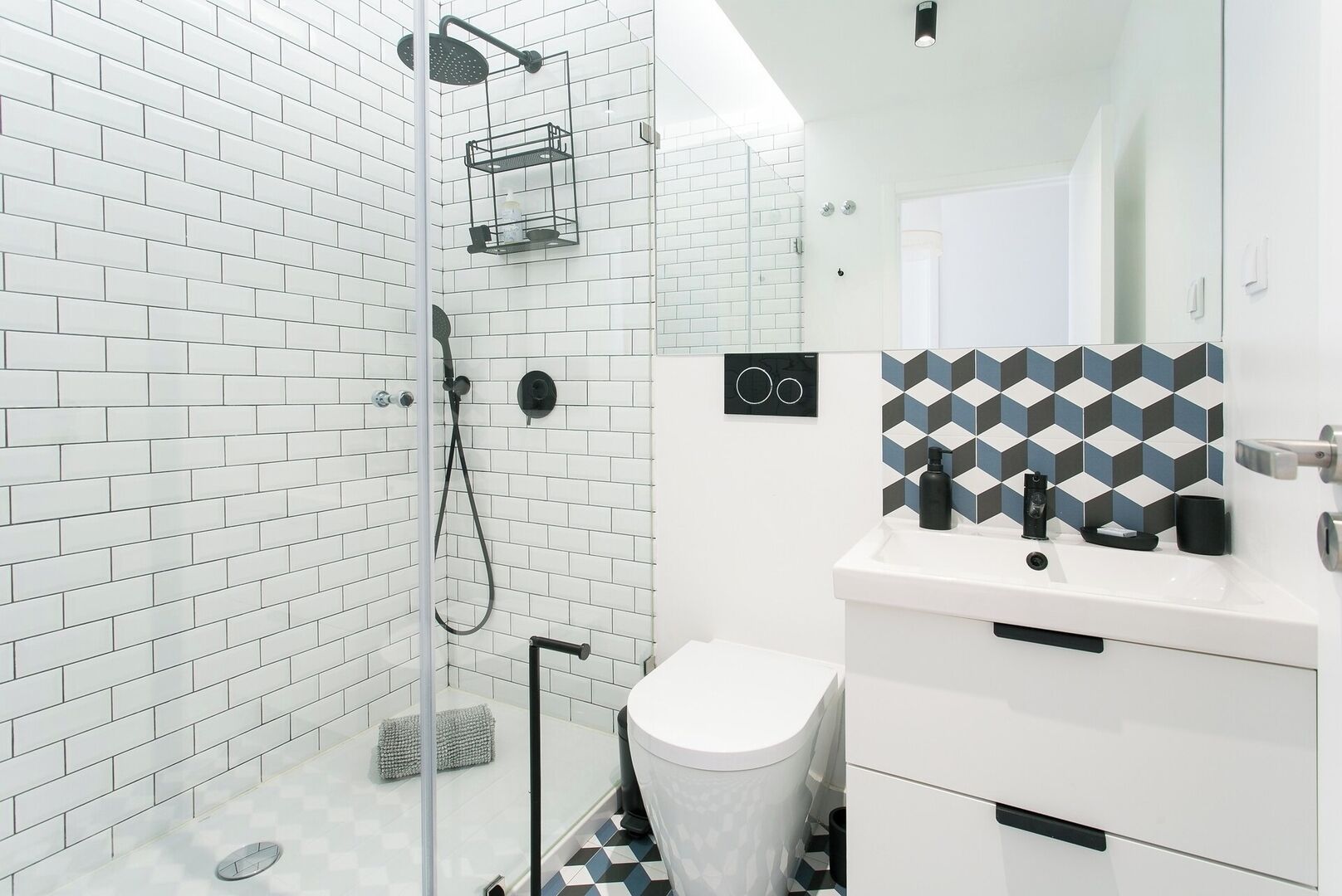 Ein modernes Badezimmer mit weißen Fliesen im U-Bahn-Stil, einer Glasdusche, einem weißen Waschbecken mit schwarzem Seifenspender und einer geometrisch gemusterten Spritzschutzeinrichtung.