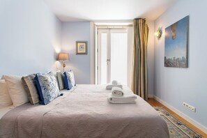 Una cama bien tendida con una colcha estampada, una lámpara de noche y un cuadro en la pared.