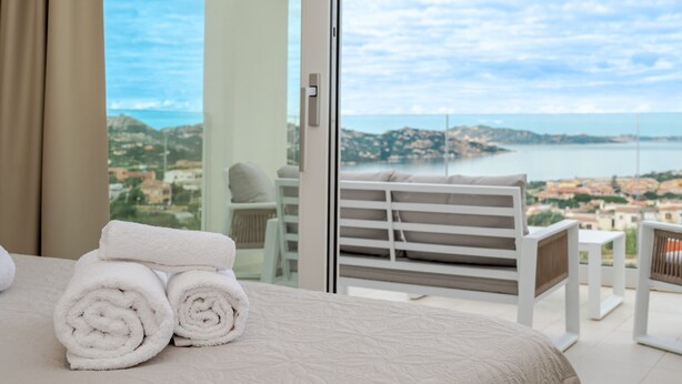 Een hotelkamer met een bed, uitzicht op zee en een balkon met een stoel en tafel.