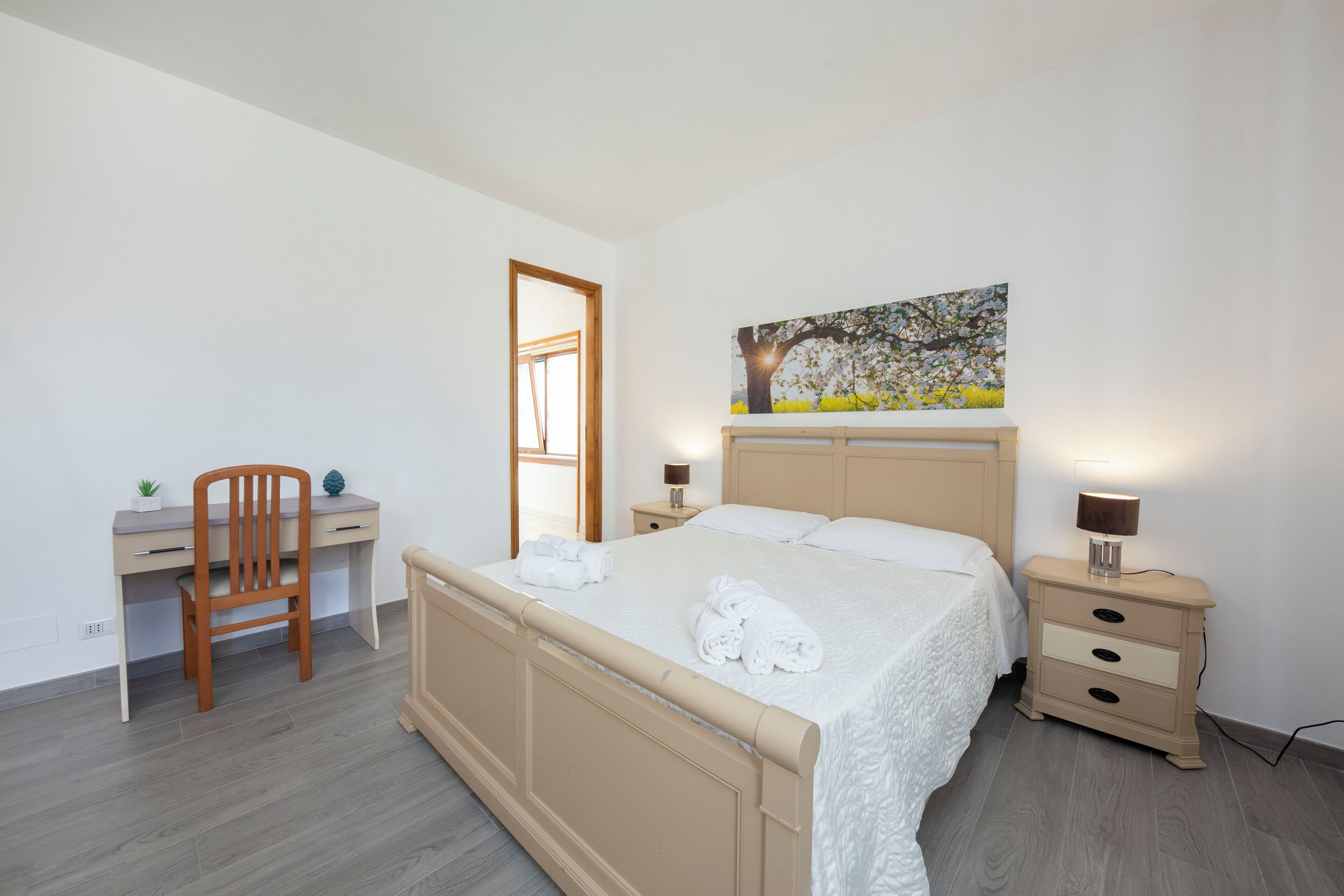 Een slaapkamer met een bed, een bureau, een stoel, een nachtkastje en een schilderij aan de muur.