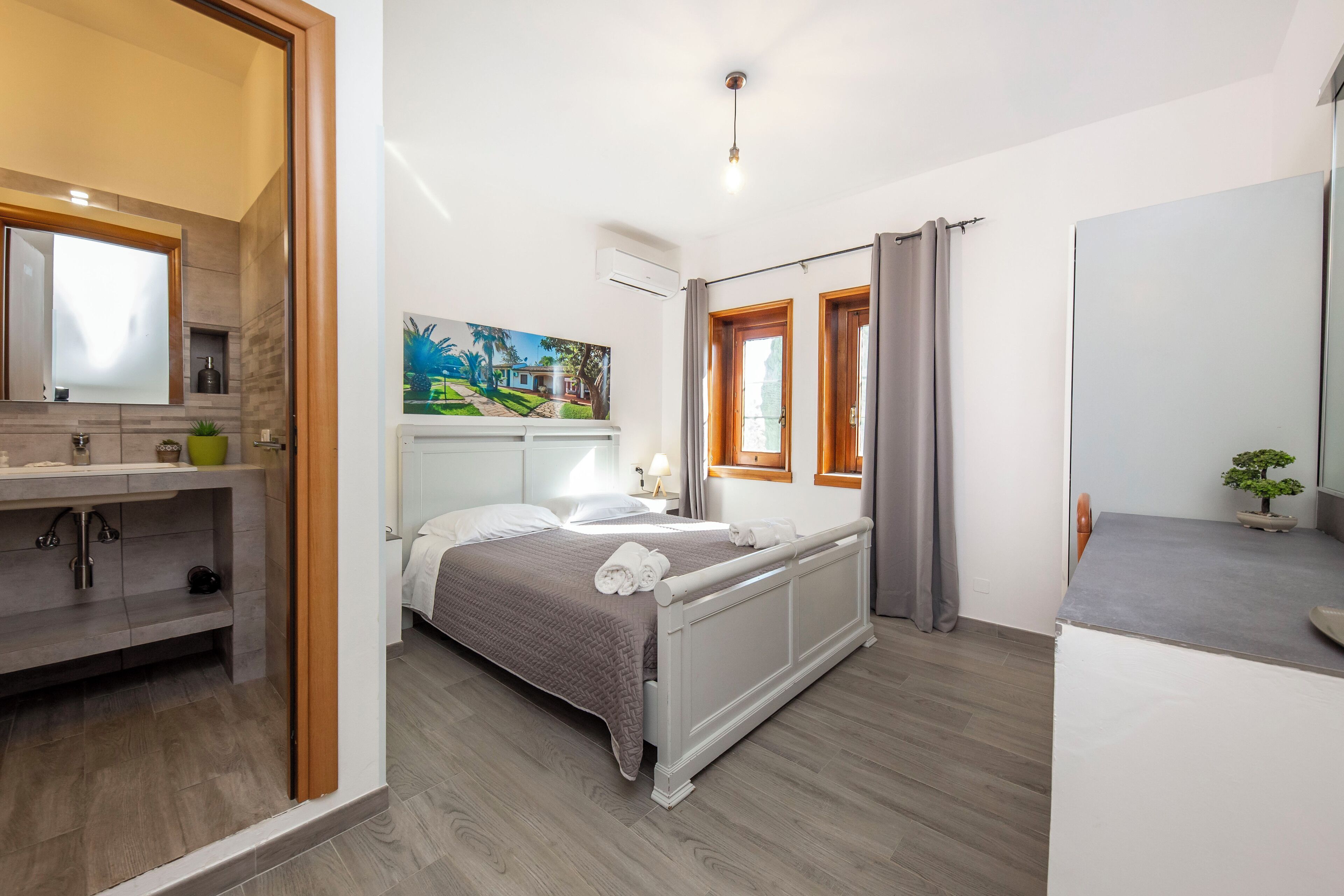 A modern bedroom with a bed, a bedside table, a mirror, a painting, and a window with curtains.