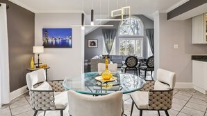 Un área de comedor con una mesa de vidrio rodeada de sillas, una lámpara colgante y un cuadro con una vista urbana en la pared.