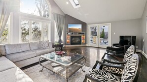 Amplia sala de estar con sofá, sillón y mesa de centro. Se observa una chimenea al fondo, y hay ventanas grandes que dejan entrar luz natural.