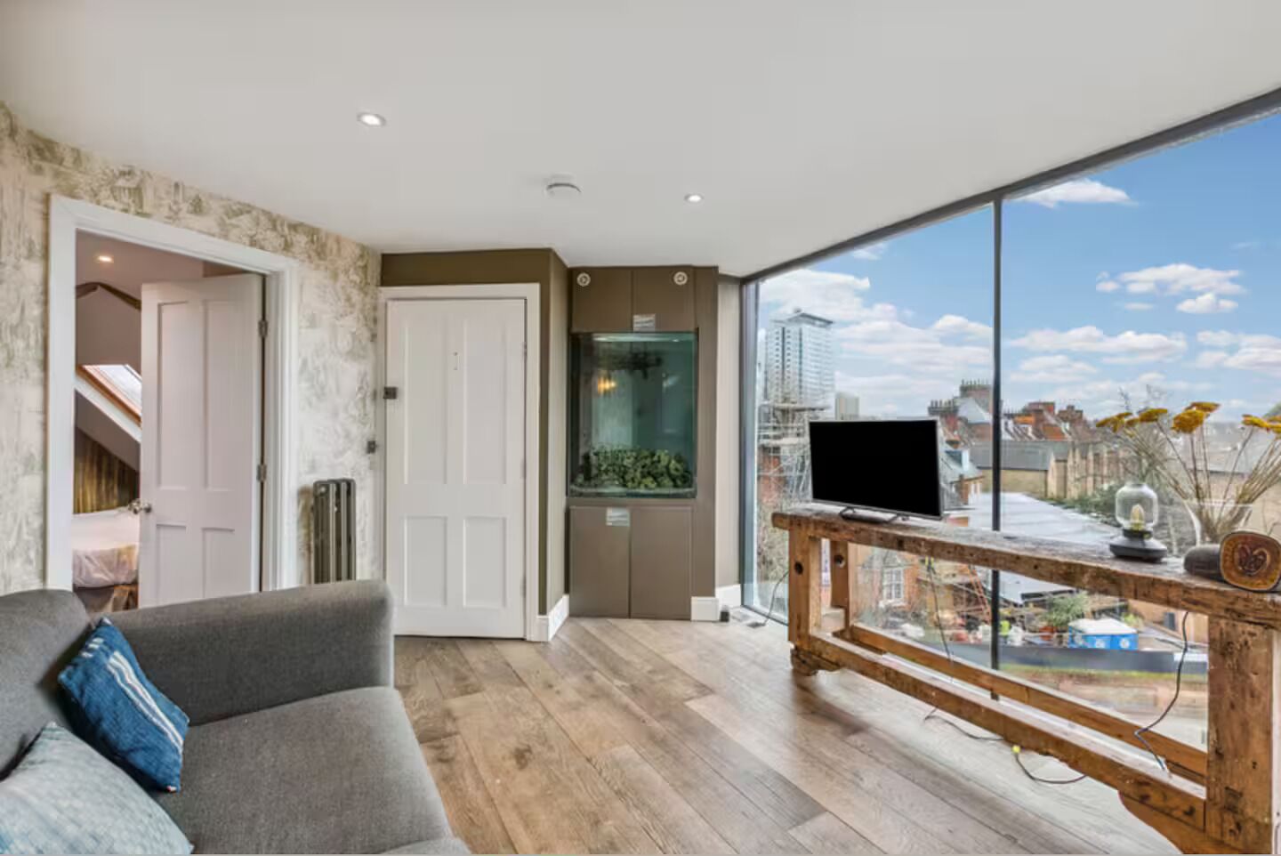 Ein modernes Wohnzimmer mit einer grauen Couch, einem Fernseher auf einem Holzboden und einem großen Fenster mit Blick auf die Stadt.