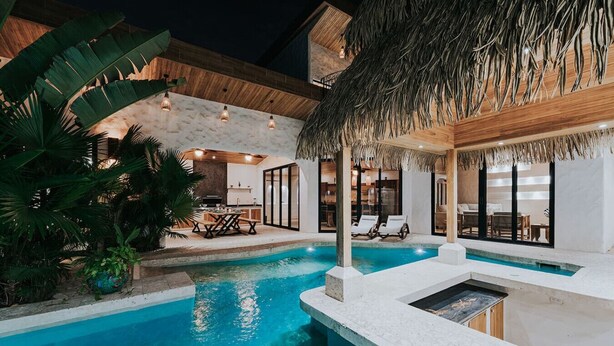 Un espace piscine doté d’une structure avec toit de chaume, de chaises longues et d’un coin repas avec une table et des chaises.