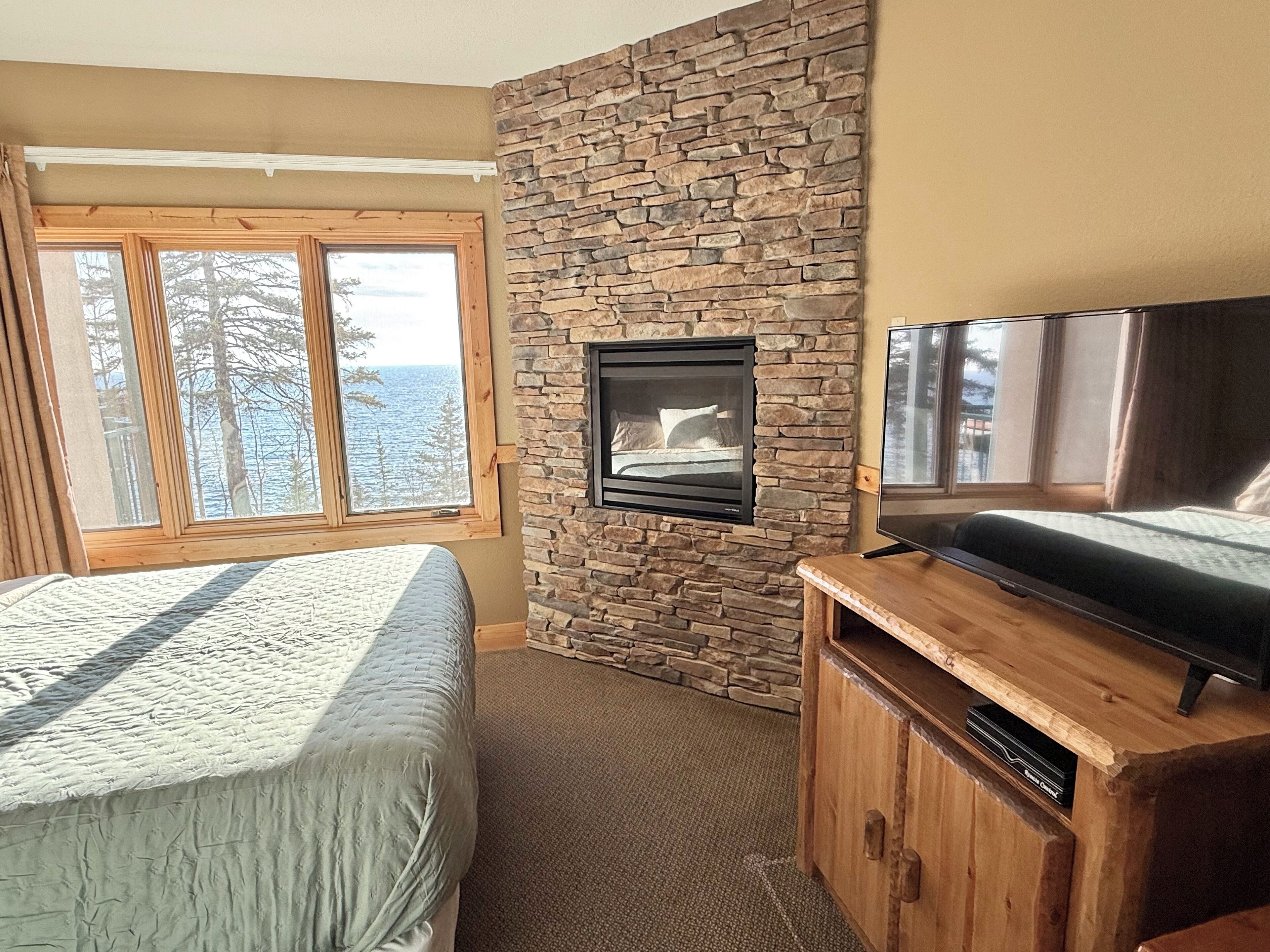 A bedroom with a stone wall, a fireplace, a bed, a TV, and a window with a view of trees and the sky.