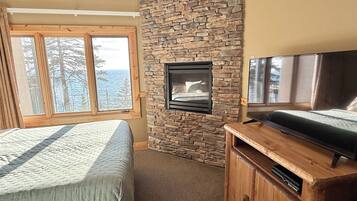 A bedroom with a stone wall, a fireplace, a bed, a TV, and a window with a view of trees and the sky.