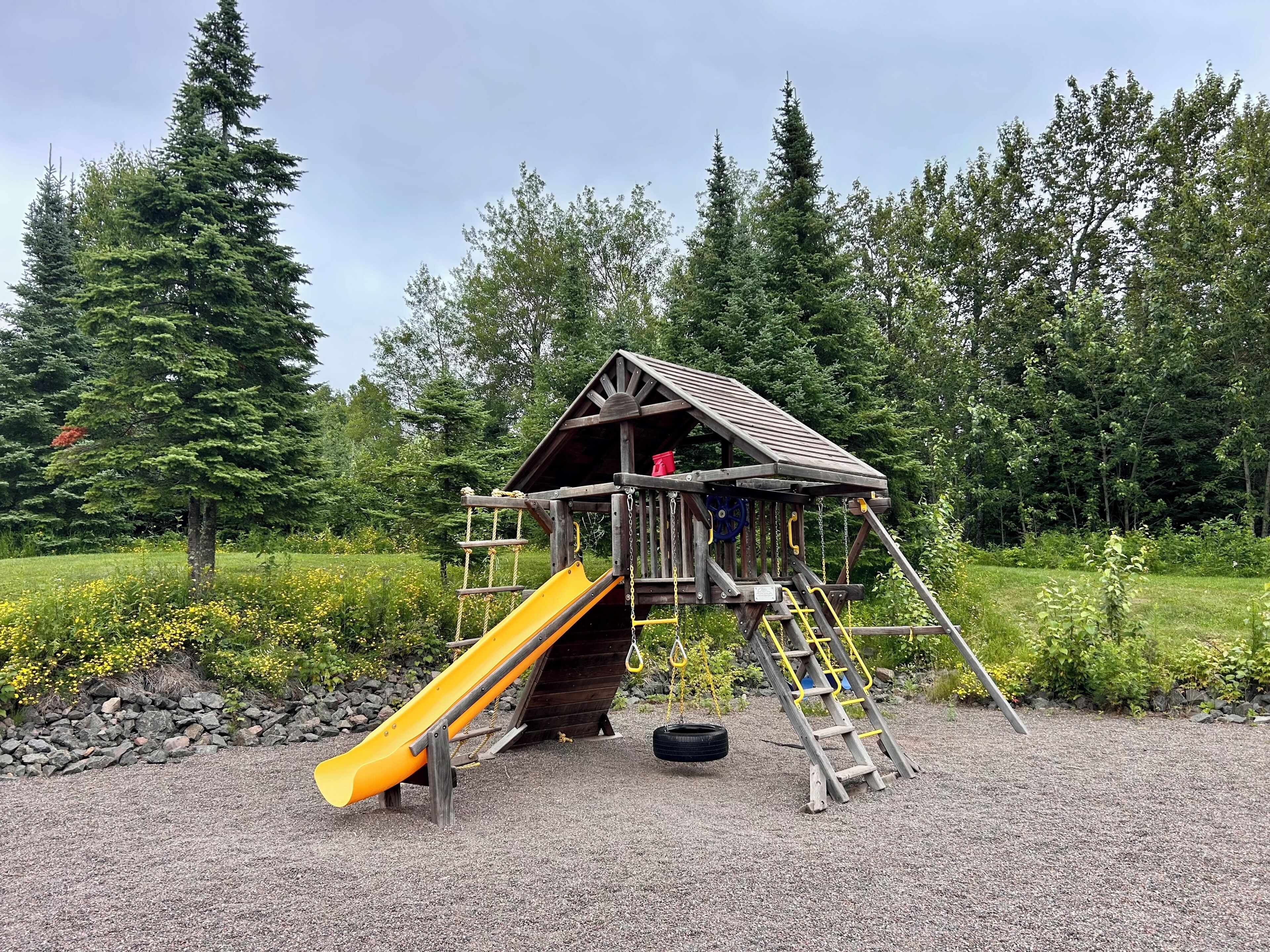 Une cabane en bois, dotée d’un toboggan et d’une échelle, entourée de gravier et de verdure.