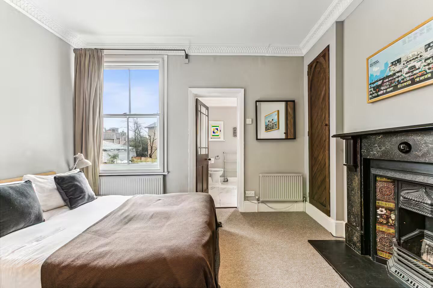 Une chambre d’hôtel avec un lit, une cheminée, une fenêtre avec des rideaux, un radiateur et un tableau encadré au mur.