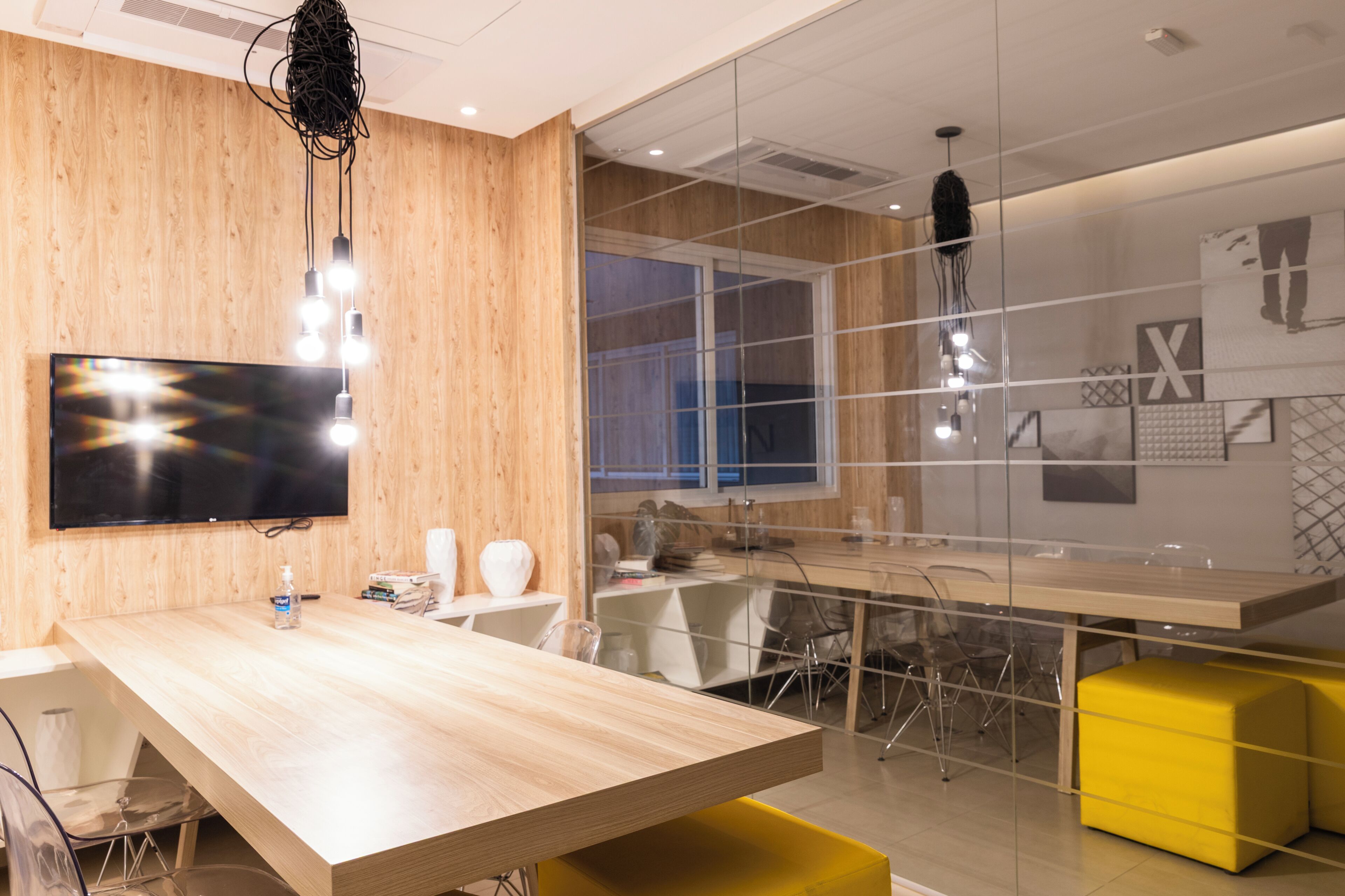 A modern meeting room with a wooden wall, a large flat-screen TV, a glass partition, and a yellow accent chair.