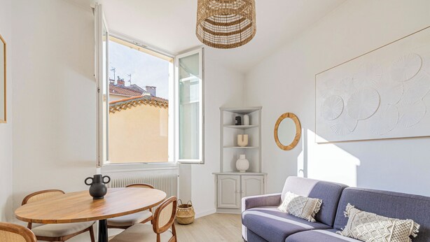 Ein modernes Wohnzimmer mit einem runden Holztisch, zwei Stühlen, einem grauen Sofa und einem großen Fenster mit Blick auf Gebäude.