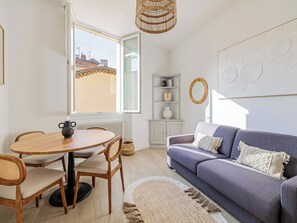Una sala de estar moderna con una mesa redonda de madera, dos sillas, un sofá gris y un ventanal con vistas a edificios.