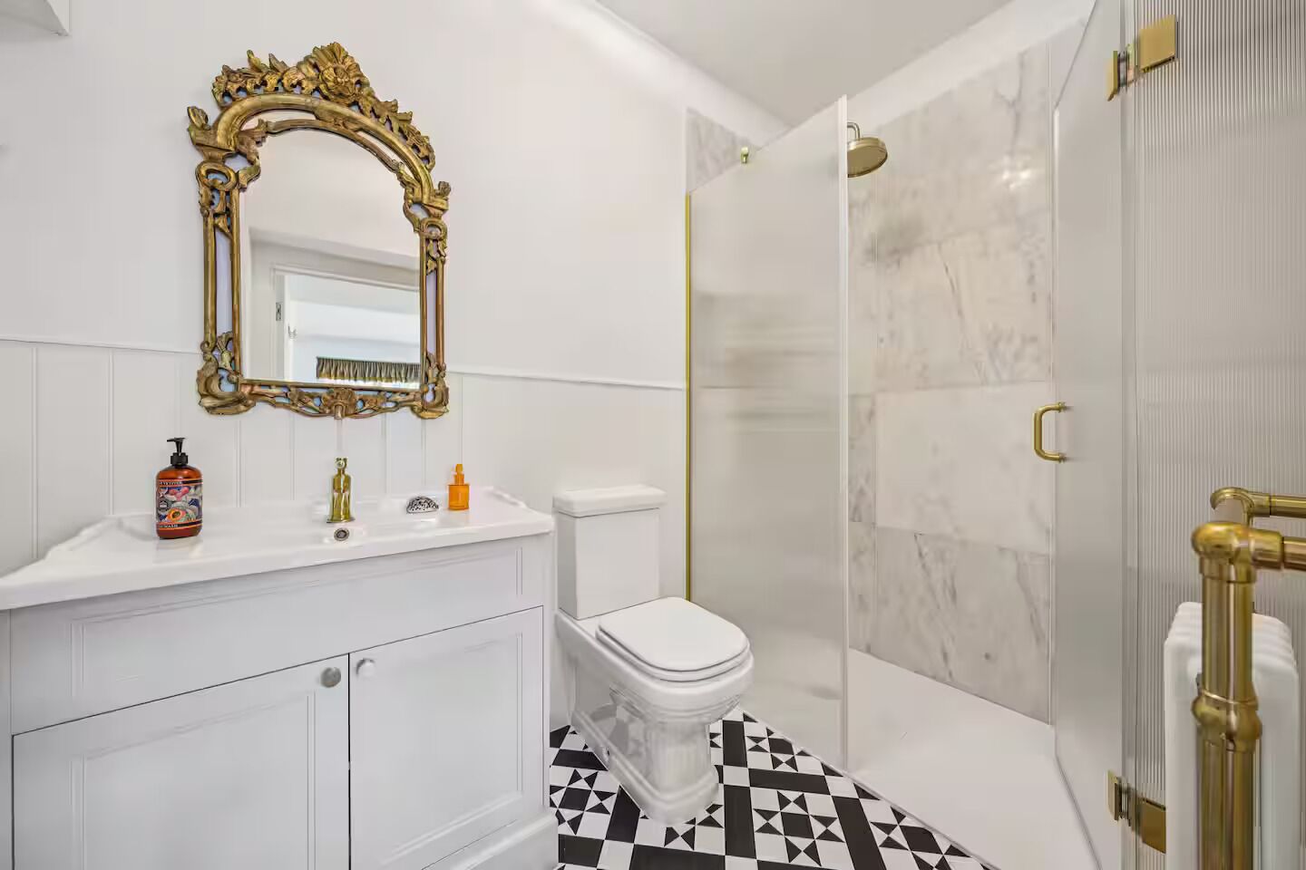 Une salle de bain avec un meuble-lavabo blanc, un miroir encadré d’or, des toilettes et une douche dotée d’une porte en verre.