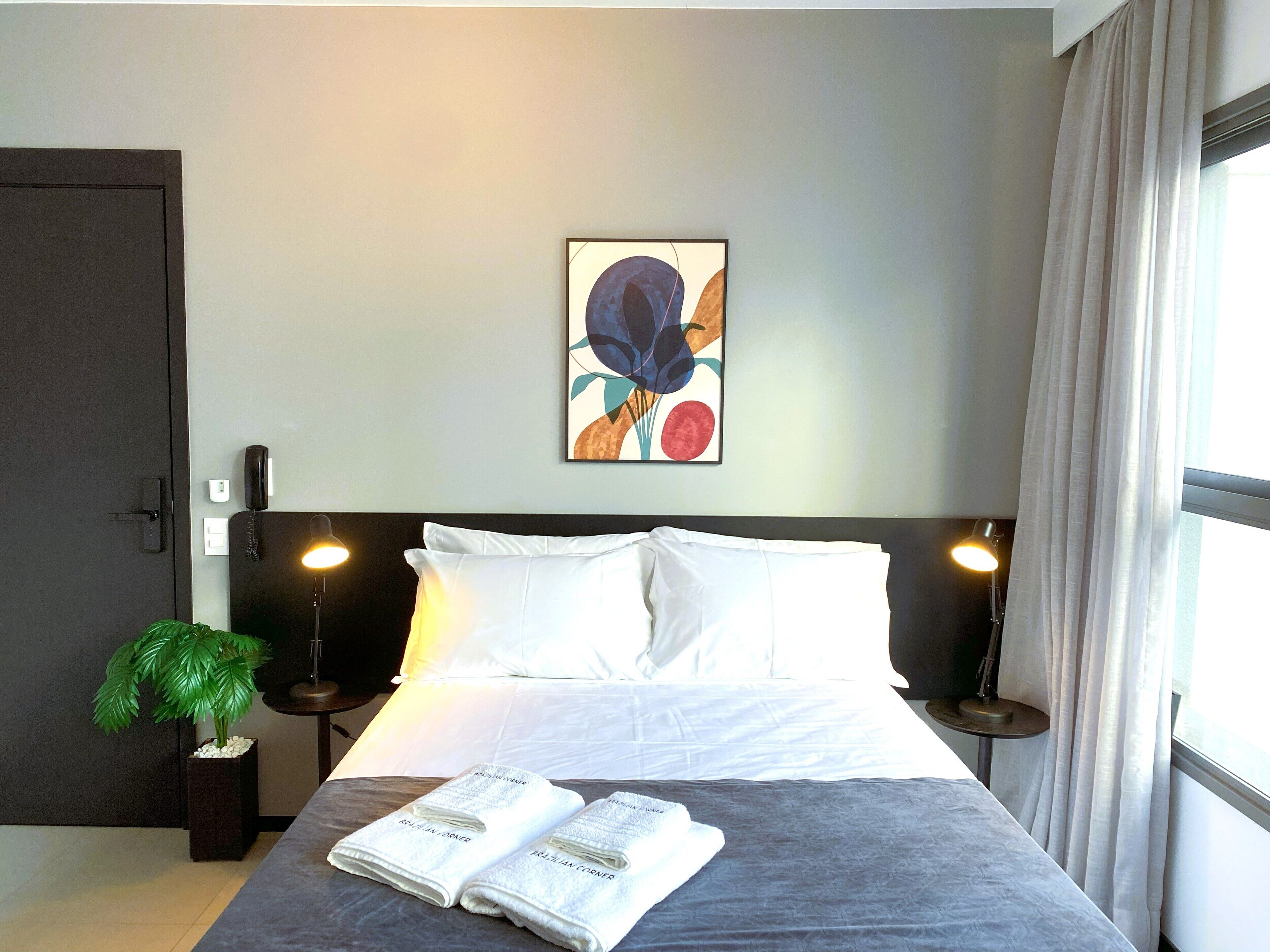 A neatly made bed with white linens, a potted plant, and a painting on the wall.
